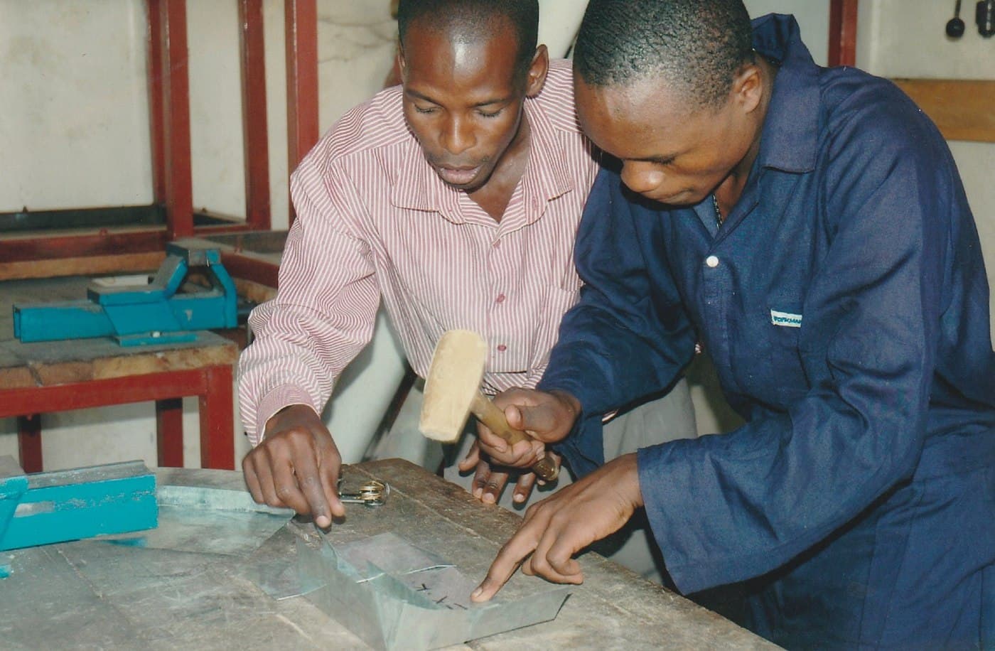 Two trainees work together filing a metal piece secured to a wall-mounted vice.
